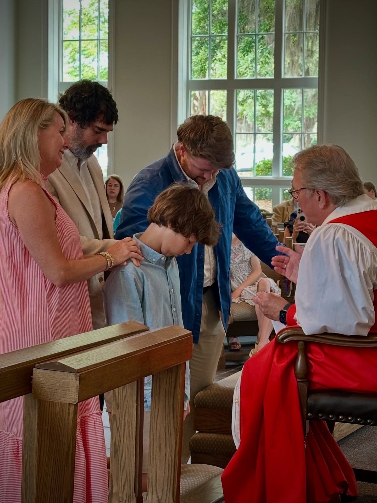 A family gathers around a young man as the bishop, in red vestments, prays a blessing over him