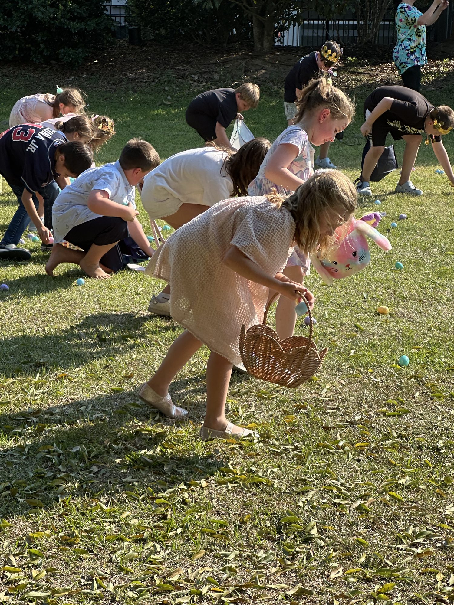 Children bending to gather colorful plastic eggs on the lawn during an Easter egg hunt, one carrying a wicker basket
