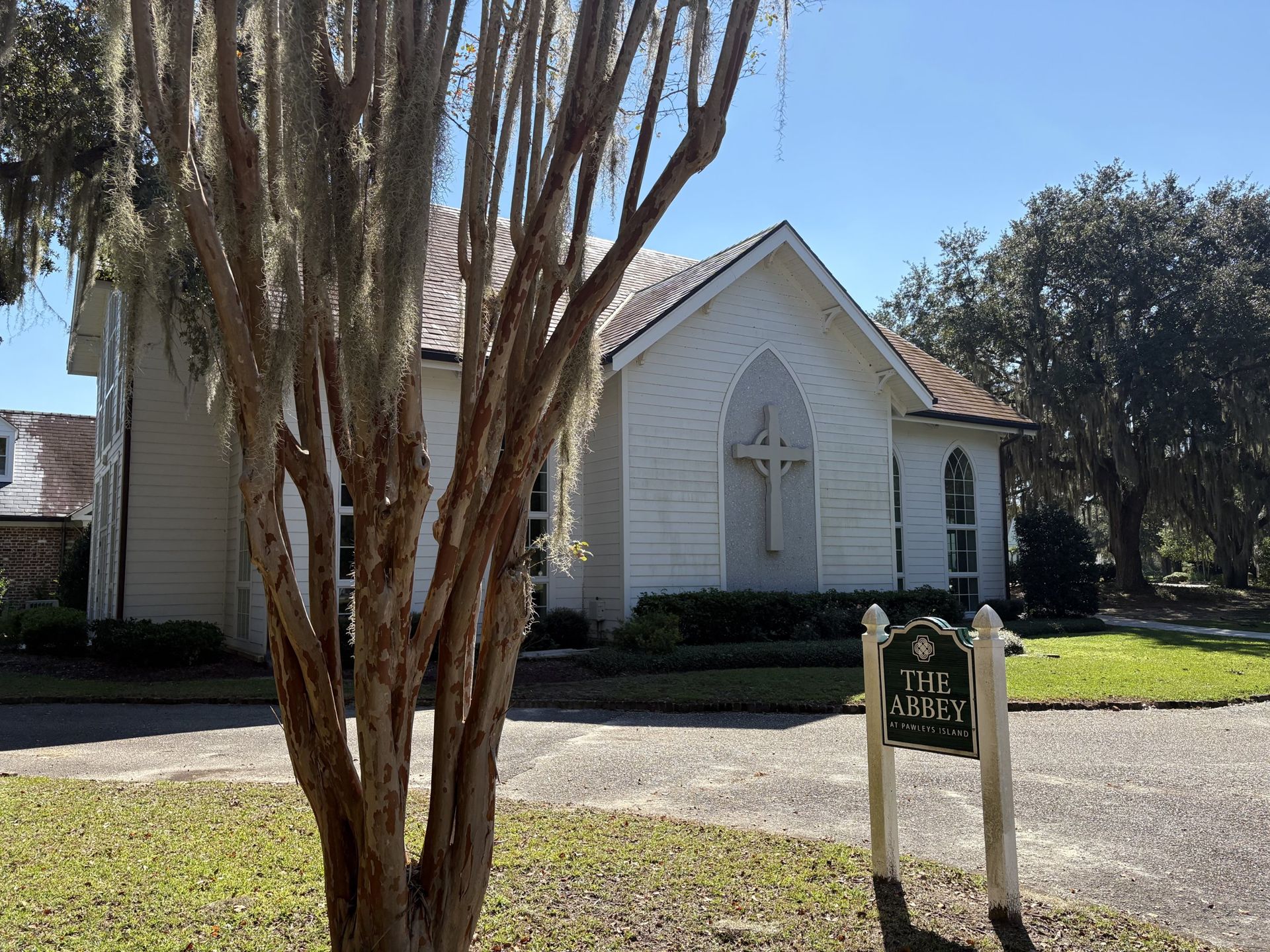 The white chapel with its stone cross and The Abbey sign, framed by a crape myrtle and live oaks in Spanish moss