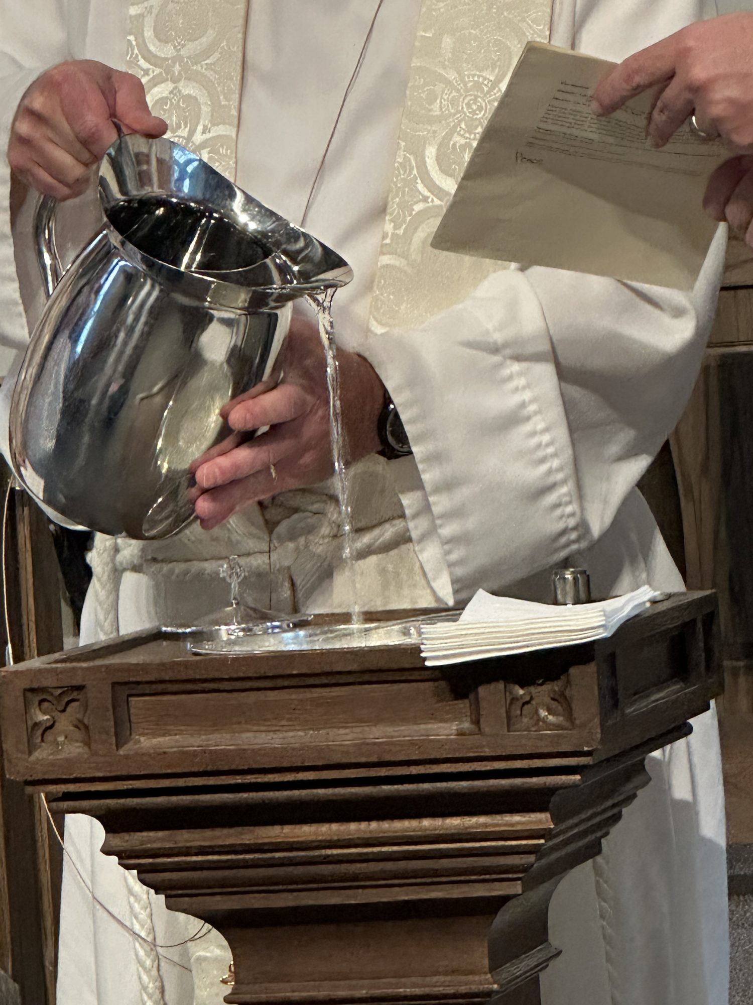 Water poured from a silver pitcher into the baptismal font by a priest in a white robe and stole